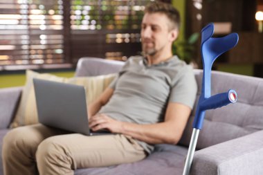 Evdeki koltukta dizüstü bilgisayar kullanan adam, dirsek değneğine odaklan.