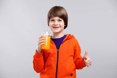 Bir bardak taze portakal suyu olan çocuk açık gri arka planda baş parmağını gösteriyor.