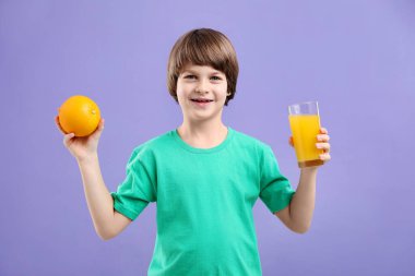 Menekşe arka planında bir bardak taze portakal suyu ve meyvesi olan çocuk.