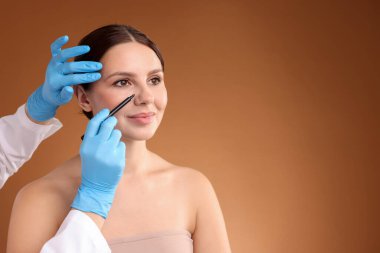 Doktor, kahverengi arkaplana karşı estetik ameliyat için hastanın yüzüne işaretler çiziyor, yakın plan. Metin için boşluk