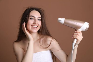 Kahverengi arka planda saç kurutma makinesiyle saçını kurutan gülümseyen kadın.