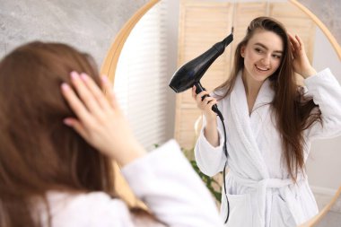 Bornozlu gülümseyen kadın banyoda aynanın yanında saç kurutma makinesiyle saçını kurutuyor.