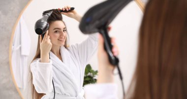 Bornozlu gülümseyen kadın saçını saç kurutma makinesiyle şekillendiriyor ve banyoda aynanın yanında tarıyor.