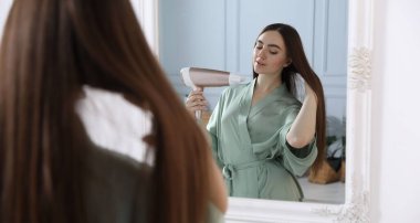 Bornozlu güzel bir kadın banyoda saçını saç kurutma makinesiyle kurutuyor.