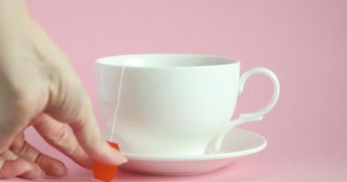 Elinde taze demlenmiş çay fincanı ve pembe arka planda beyaz çay poşeti olan bir kadın, yakın plan.