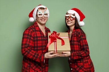 Noel Baba şapkalı ve pijamalı mutlu çift. Gri yeşil arka planda bir Noel hediyesi.
