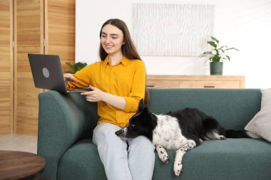 Evdeki kanepede dizüstü bilgisayarı ve sevimli köpeğiyle gülümseyen kadın.