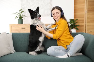 Evdeki kanepede sevimli köpeğiyle kulaklıklı, gülümseyen bir kadın.