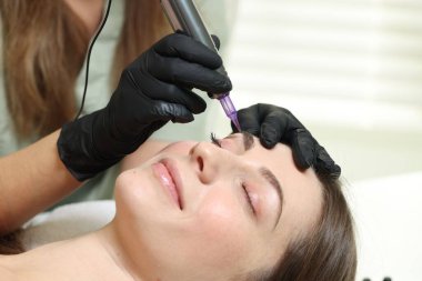 Güzellik uzmanı, kuaförde genç bir kadına kalıcı kaş makyajı yapıyor.