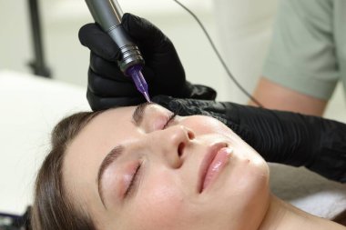 Güzellik uzmanı, içeride genç bir kadına kalıcı kaş makyajı yapıyor.