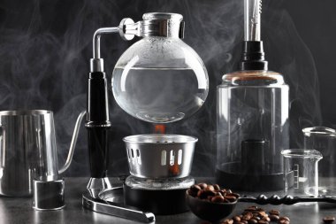 Süpürge kahve makinesi, kahve ve fasulye yapmak için diğer ekipmanlar gri masada, siyah arka planda dumanlı, yakın plan