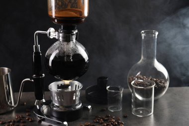 Vakumlu kahve makinesinde aromatik kahve demlemek, laboratuvar bardağı ve fasulye gri masada koyu arkaplanda dumanla kaplanmak, yakın plan