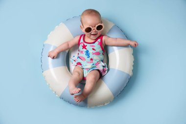 Şişirilebilir halkada güneş gözlüklü şirin bir bebek açık mavi arka planda, üst tarafta.