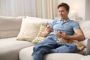Evdeki kanepede akıllı telefon kullanan sevimli kedisiyle gülümseyen bir adam. Metin için boşluk