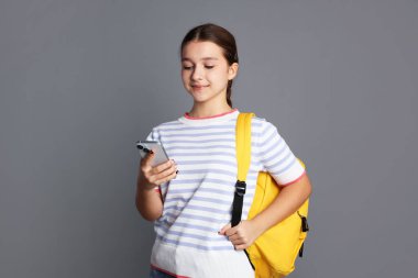 Sırt çantalı genç kız gri arka planda akıllı telefon kullanıyor.