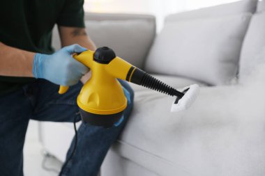 Haşere kontrol işçisi kanepeyi temizliyor. Kapalı alanda buhar temizleyicisi var.