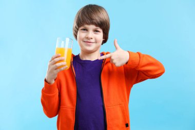 Açık mavi arka planda taze portakal suyu bardağını gösteren çocuk.