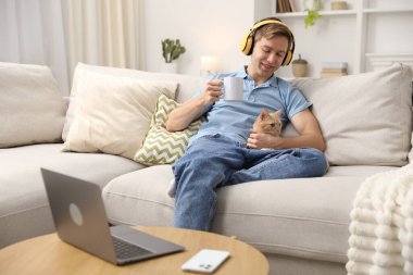 Evde sevimli kedisi ve kupasıyla kulaklıklı gülümseyen bir adam.