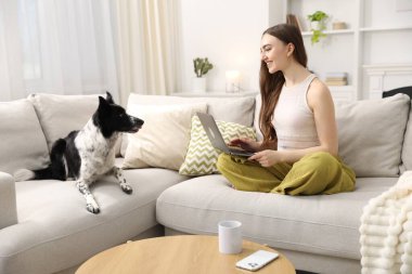 Evdeki kanepede dizüstü bilgisayarı ve sevimli köpeğiyle gülümseyen kadın.