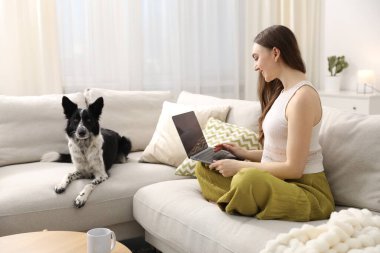 Evdeki kanepede dizüstü bilgisayarı ve sevimli köpeğiyle gülümseyen kadın.