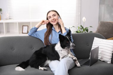Akıllı telefonuyla gülümseyen bir kadın ve evdeki kanepede oturan sevimli köpeği.