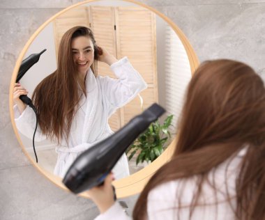 Bornozlu gülümseyen kadın banyoda aynanın yanında saç kurutma makinesiyle saçını kurutuyor.