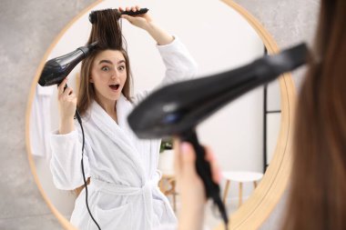 Bornozlu duygusal bir kadın saçını saç kurutma makinesiyle şekillendiriyor ve banyoda aynanın yanında tarıyor.