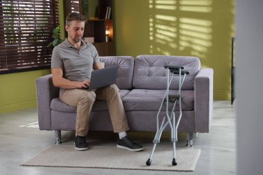 Evdeki koltuk değneklerinin yanındaki koltukta dizüstü bilgisayar kullanan bir adam.