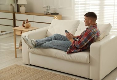 Akıllı telefonlu genç adam evdeki kanepede dinleniyor.