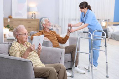 Huzurevinde kitap okuyan yaşlı erkek ve kadına bir bardak su veren bakım işçisi.