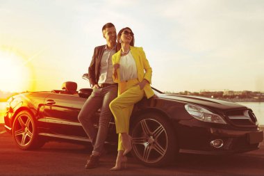 Güneşli bir günde üstü açık lüks bir arabanın yanında şık bir çift.