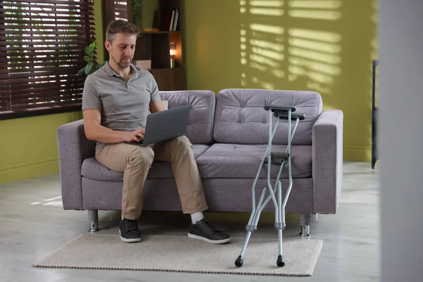 Evdeki koltuk değneklerinin yanındaki koltukta dizüstü bilgisayar kullanan bir adam.