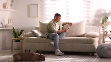 Akıllı telefonlu genç adam evdeki şirin köpeğin yanındaki kanepede dinleniyor.