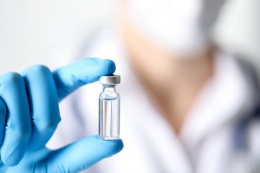 Hemşire arka planda aşı şişesi tutuyor, yakın plan.