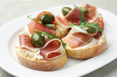 Krem peynirli lezzetli bruschettalar, prosciutto, yeşil zeytin ve roka gri masada, yakın plan.