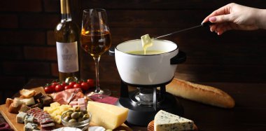 Kadın, ahşap masada erimiş peynirle fondü kabına (caquelon) bir parça ekmek daldırıyor.