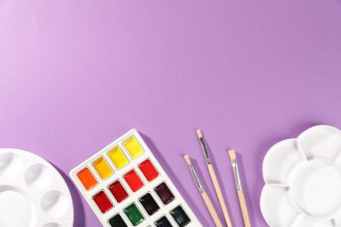 Suluboya boyalar, paletler ve fırçalar mor arka planda, düz yerde. Metin için boşluk
