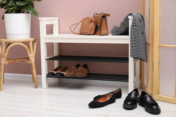 Shoe storage bench with different footwear, bags, jacket and houseplant near dusty pink wall indoors
