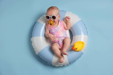 Güneş gözlüklü, oyuncak ördekli, şişme yüzüklü, açık mavi arka planda şirin bir bebek.