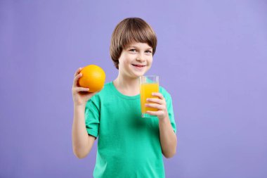 Menekşe arka planında bir bardak taze portakal suyu ve meyvesi olan çocuk.