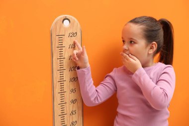 Küçük kız turuncu duvarın yanında boyunu ölçüyor.