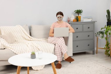 Yüzünde kilden bir maske olan bir kadın evinde dizüstü bilgisayar kullanıyor.