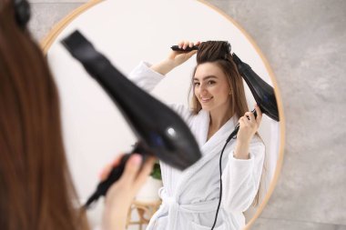 Bornozlu gülümseyen kadın saçını saç kurutma makinesiyle şekillendiriyor ve banyoda aynanın yanında tarıyor.