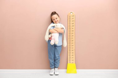 Oyuncak tavşancığı olan küçük bir kız bej duvarın yanında boyunu ölçüyor.