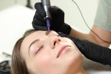 Güzellik uzmanı, içeride genç bir kadına kalıcı kaş makyajı yapıyor.