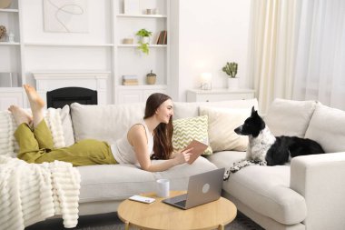 Sevimli köpeğiyle evdeki kanepede kitap okuyan gülümseyen kadın.