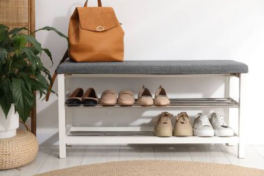 Ayakkabı bankı, farklı ayakkabılar, sırt çantası ve beyaz duvarın yanındaki ev bitkisi.