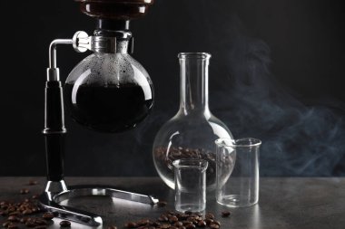 Vakumlu kahve makinesinde aromatik kahve demlemek, laboratuvar bardağı ve fasulye gri masada koyu arkaplanda dumanla kaplanmak, yakın plan