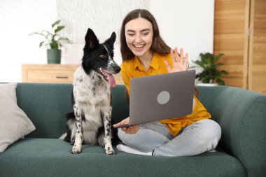Dizüstü bilgisayarlı gülümseyen kadın merhaba diyor ve evdeki kanepede sevimli köpeği oturuyor.