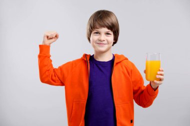 Bir bardak taze portakal suyu olan çocuk açık gri arka planda güç gösteriyor.